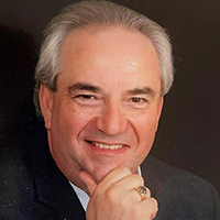 A man in a black suit sits with his right hand holding his chin