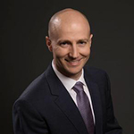 A man in a black suit, white collared shirt, and dark tie stands and smiles