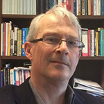 A man with glasses sits in front of a book shelf and smiles